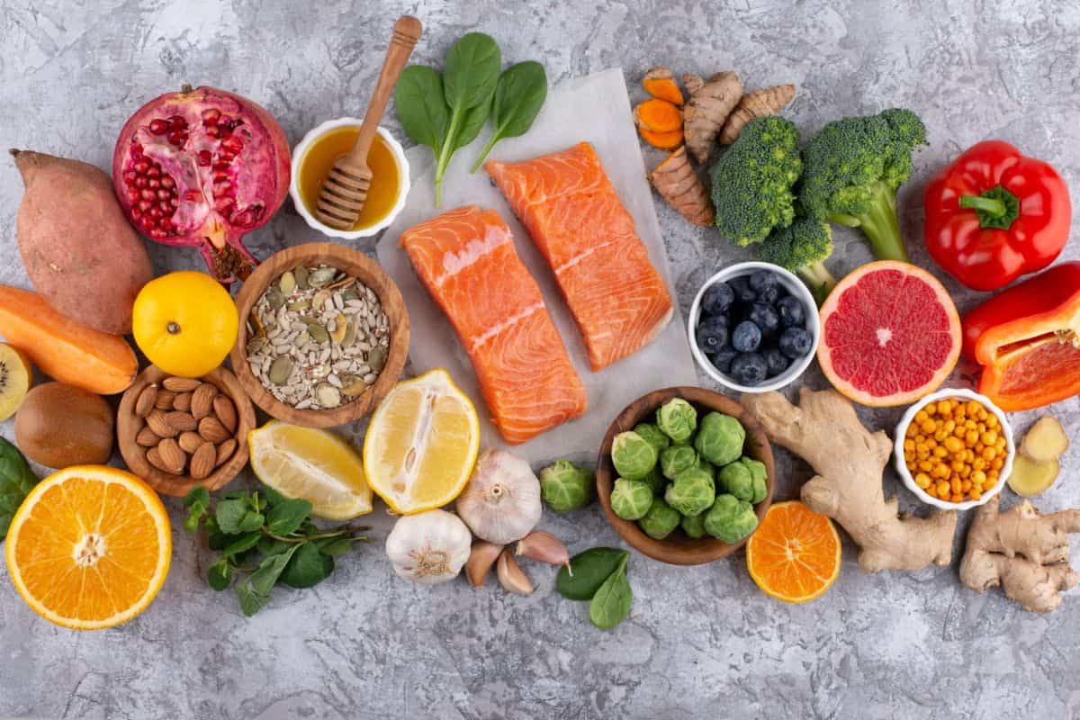 An assortment of healthy foods including salmon, fruits, vegetables, nuts, seeds, honey, and grains arranged on a textured surface.