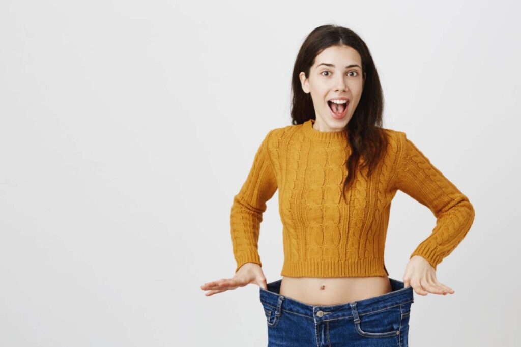 A woman in a mustard yellow sweater is happily showing off her significant weight loss by holding out the waistband of her oversized jeans.