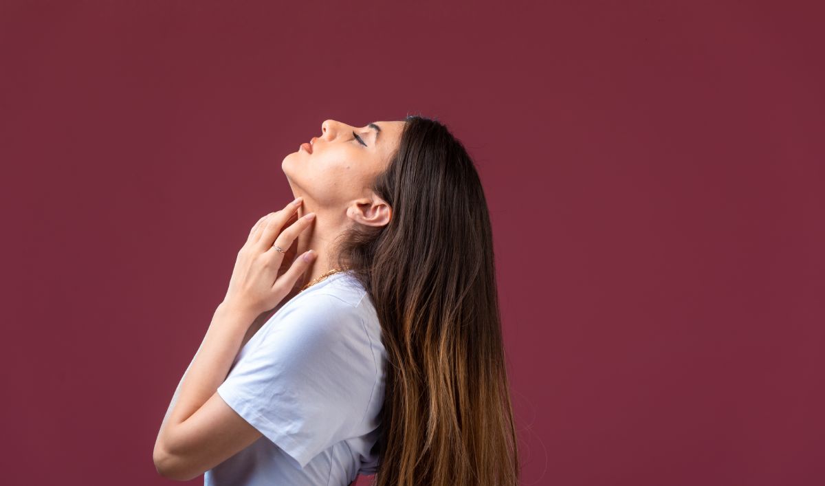 A woman with long brown hair performing a tech neck exercise. She is tilting her head back, stretching her neck while gently touching her throat. The background is a solid maroon color.