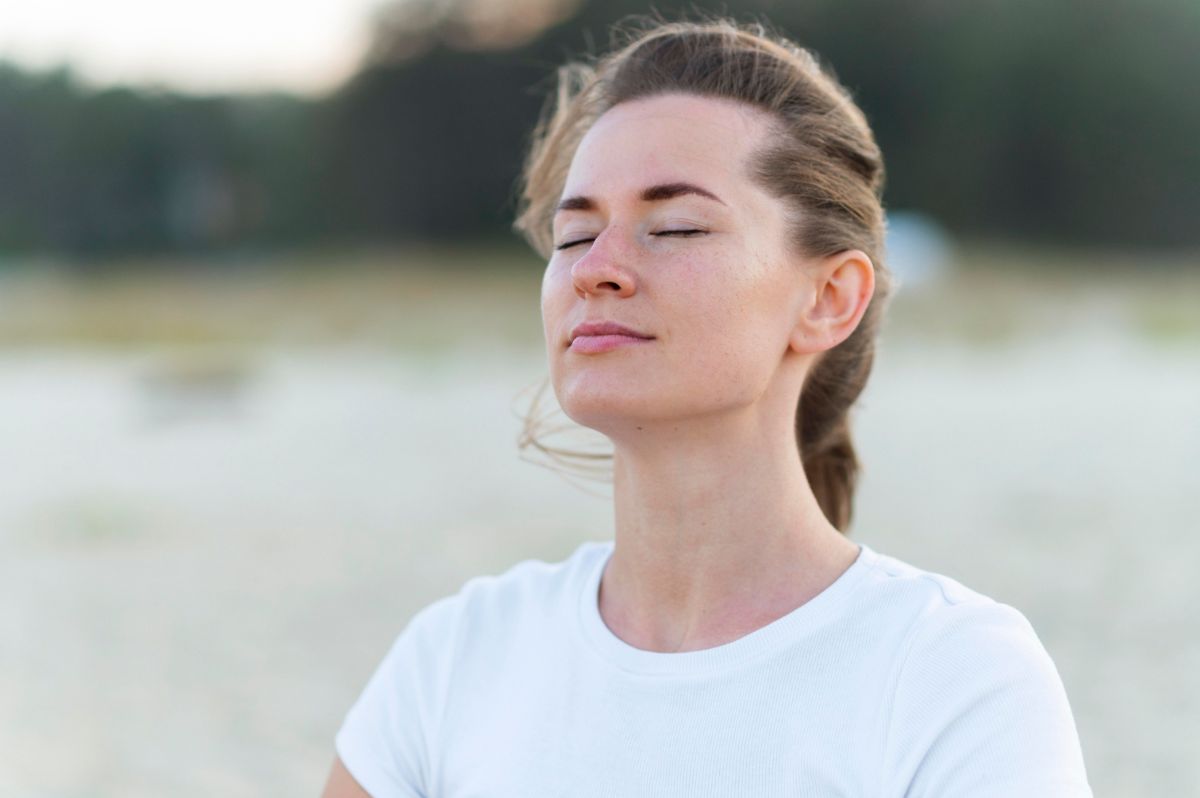 A young woman with closed eyes stands outdoors, appearing relaxed and serene. She is wearing a white T-shirt and her hair is tied back. The background is blurred, suggesting a natural, possibly beach setting.