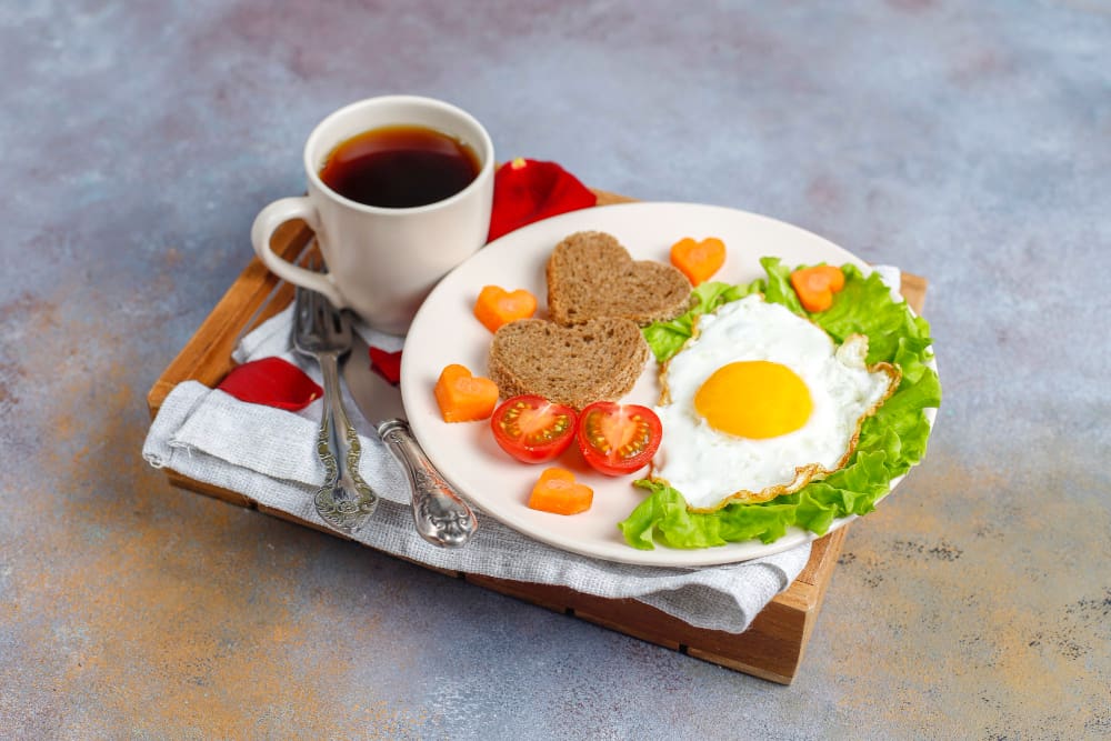 A breakfast plate featuring a sunny-side-up egg on a bed of lettuce, heart-shaped pieces of toast, cherry tomato halves, and small heart-shaped carrot pieces, accompanied by a cup of black coffee. The setup is on a wooden tray with a white cloth and cutlery.