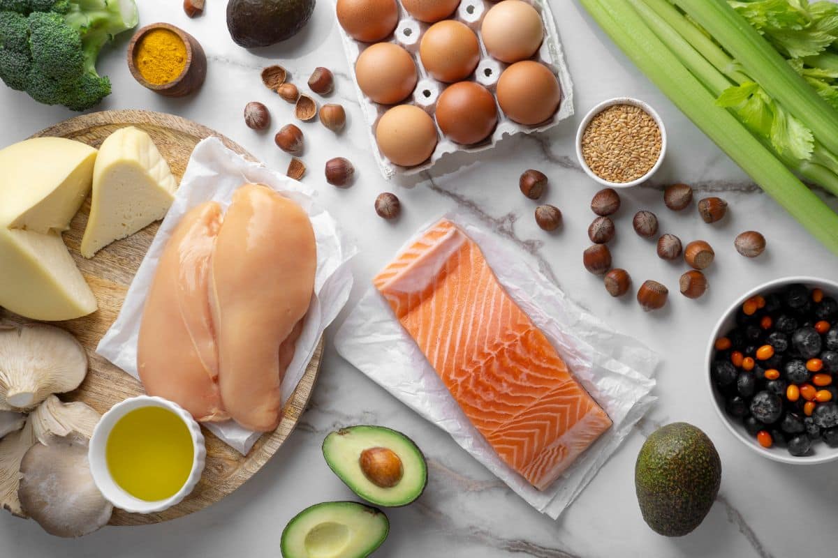 A variety of healthy foods including chicken breasts, a salmon fillet, eggs, avocados, nuts, celery, cheese, blueberries, and oil displayed on a marble countertop.