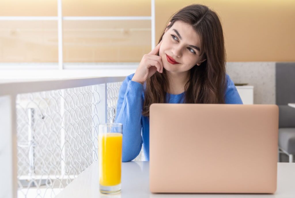 Woman pondering while looking at a laptop with a glass of orange juice beside her, illustrating the question under the subheading: How Long Does It Take for Different Vitamins to Start Working?