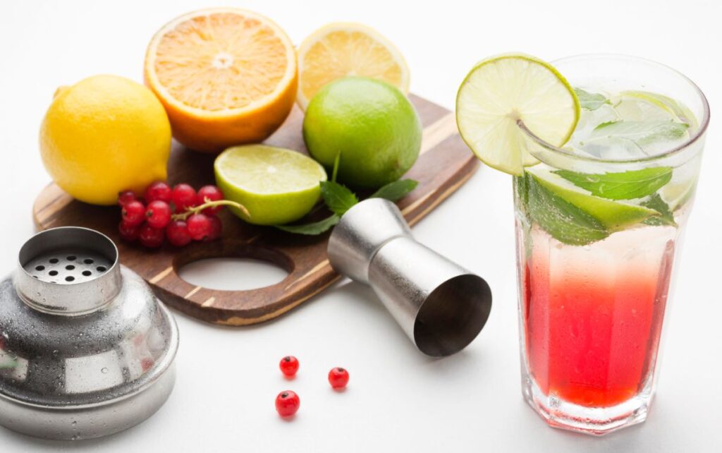 A glass of a colorful drink with lime slices and mint, surrounded by various fresh fruits including lemon, orange, lime, and red berries, with a metal cocktail shaker and jigger on the side.