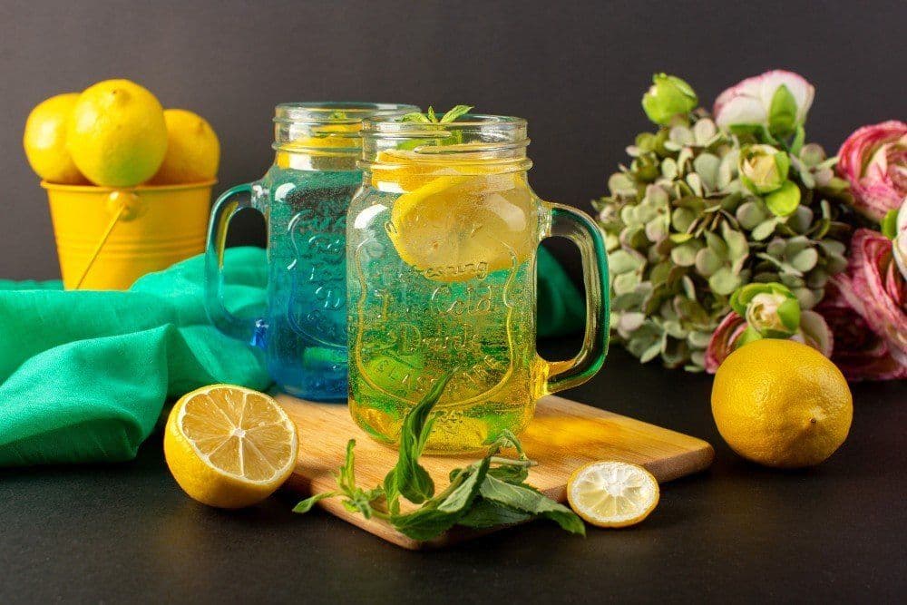 Two mason jars filled with refreshing lemon-infused water, garnished with lemon slices and mint leaves. A whole lemon and a halved lemon are placed in the foreground. In the background, a yellow bucket filled with lemons, a green cloth, and a bouquet of flowers are visible.