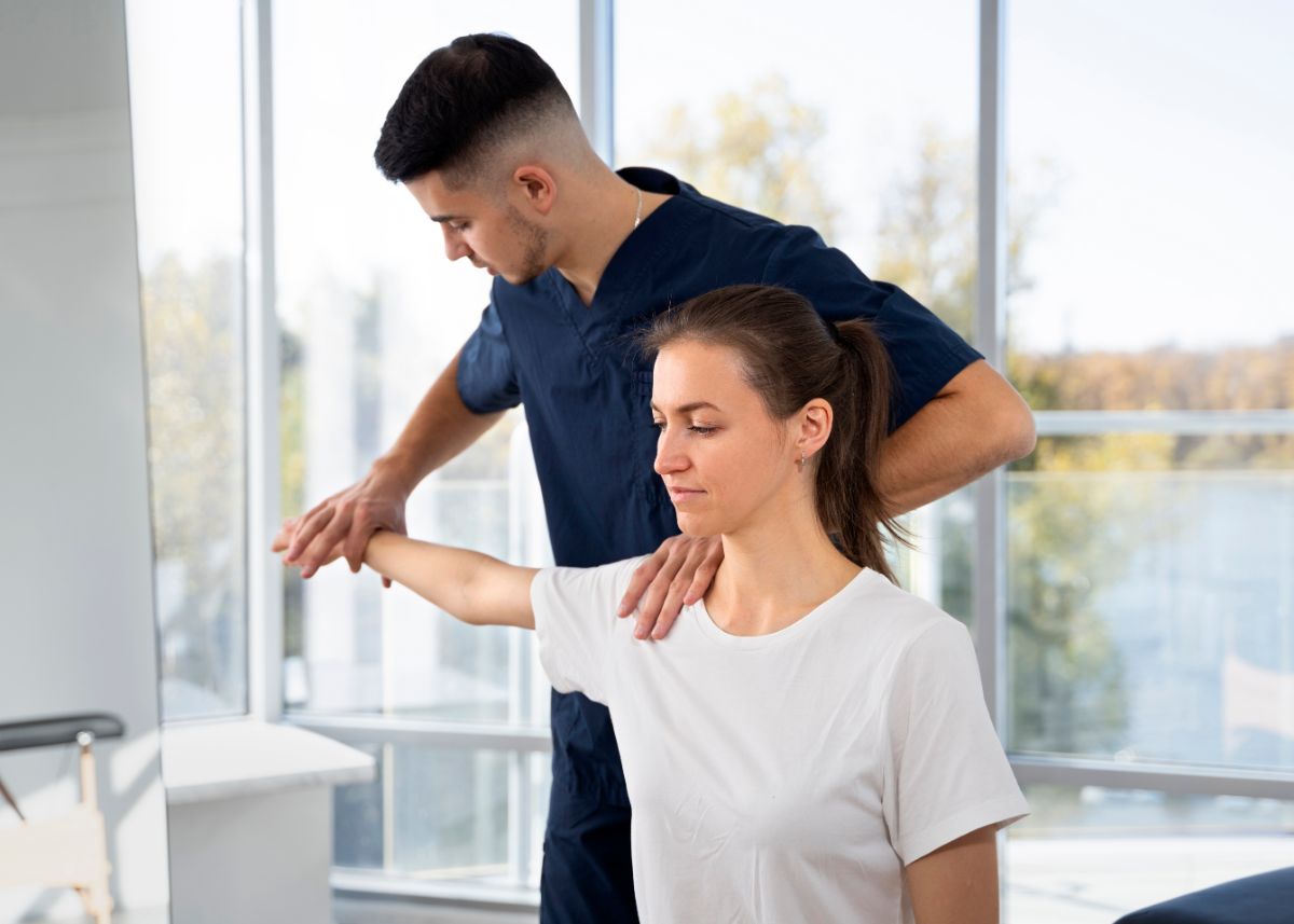 A physical therapist assists a woman in performing shoulder and neck exercises in a bright, sunlit room. The woman is practicing tech neck exercises to alleviate neck strain.