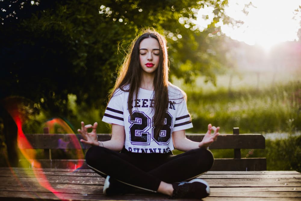 A young woman with long hair sits cross-legged on a wooden platform outdoors, meditating with her eyes closed. She is wearing a white T-shirt with the number 28 and black leggings. The background is filled with lush greenery and sunlight filtering through the trees.