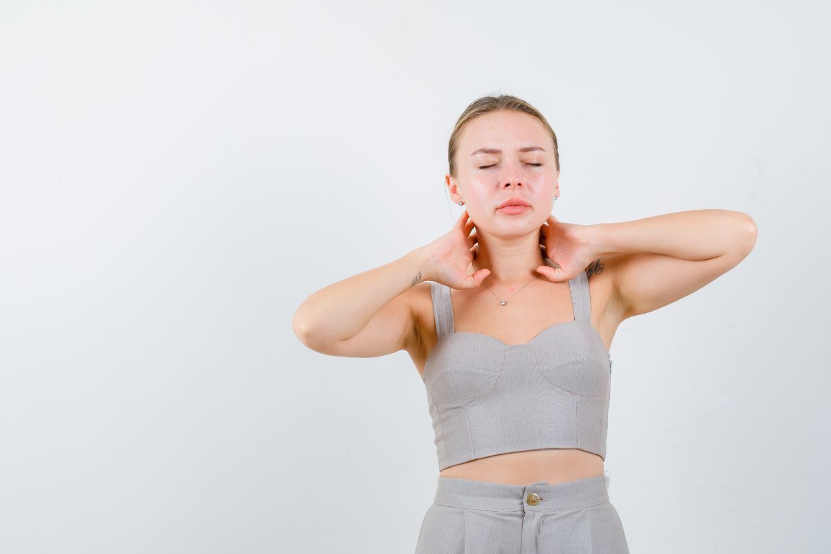 A woman with blonde hair performing a tech neck exercise. She is massaging her neck with both hands, eyes closed, and wearing a light grey outfit. The background is a plain white wall.