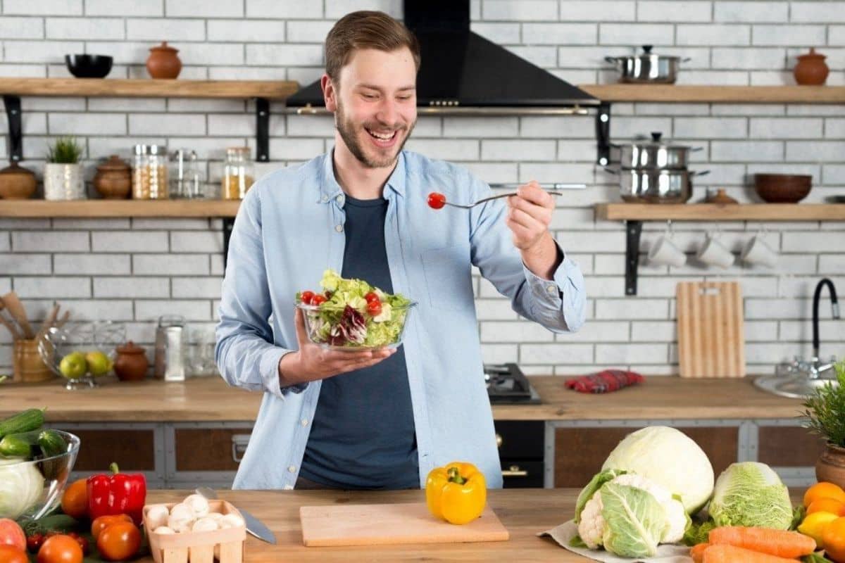 A man holding a bowl of salad, smiling and using chopsticks to pick up a cherry tomato in a kitchen filled with various fresh vegetables.