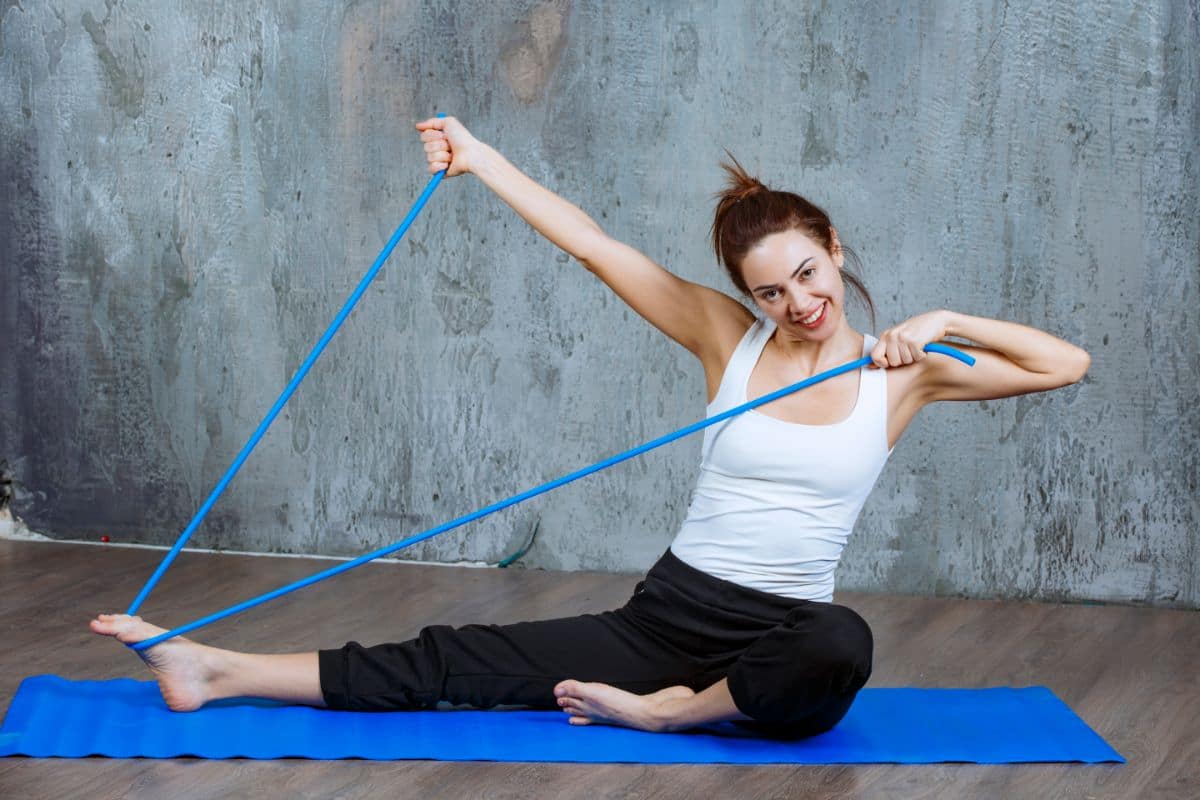 A woman performing a stretching exercise on a blue yoga mat using a resistance band. She is dressed in a white tank top and black pants, with her hair tied up in a bun. The background is a textured grey wall.