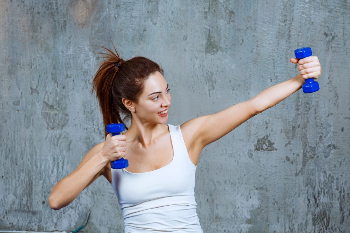 A woman in a white tank top performs a punch exercise with small blue dumbbells against a concrete wall background. This exercise is part of her tech neck exercises routine.