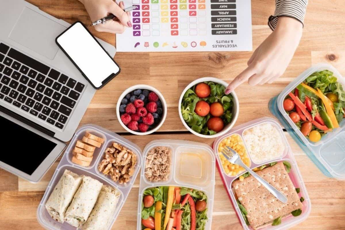 A top-down view of a table with various meal prep containers filled with healthy foods, a laptop, a smartphone, and a meal planning chart. Two hands are seen pointing and writing on the chart. The foods include salads, wraps, berries, nuts, and other nutritious items.