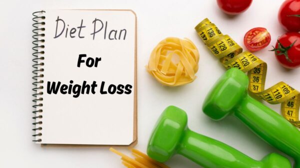 An open notebook with "The Menopause Diet 5 Day Plan for Weight Loss" written on it, surrounded by healthy items like tomatoes, a tape measure, green dumbbells, and uncooked pasta on a white surface.