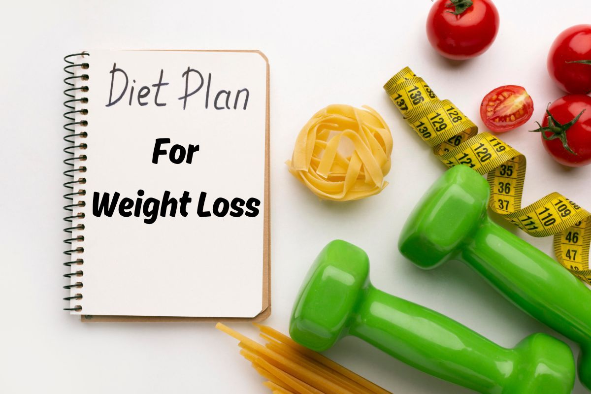 An open notebook with "The Menopause Diet 5 Day Plan for Weight Loss" written on it, surrounded by healthy items like tomatoes, a tape measure, green dumbbells, and uncooked pasta on a white surface.