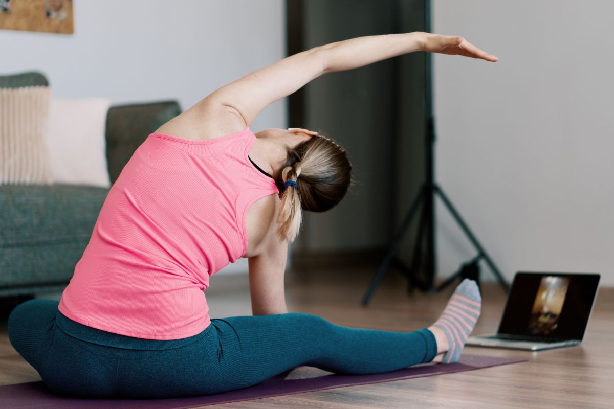 A woman in a pink tank top and blue leggings performs a side stretch on a yoga mat while watching a video on a laptop. This exercise is part of her tech neck exercises routine.