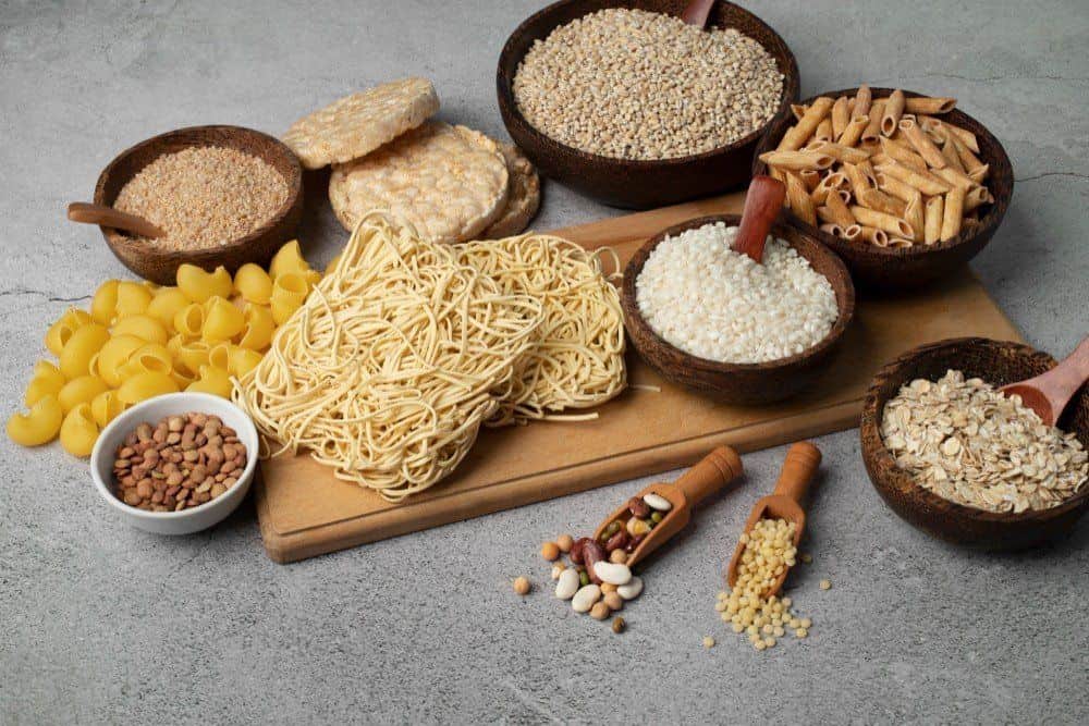 A variety of grains, legumes, and pasta arranged on a wooden board and in wooden bowls. The selection includes spaghetti, penne, shell pasta, rice cakes, oats, millet, lentils, and mixed beans.