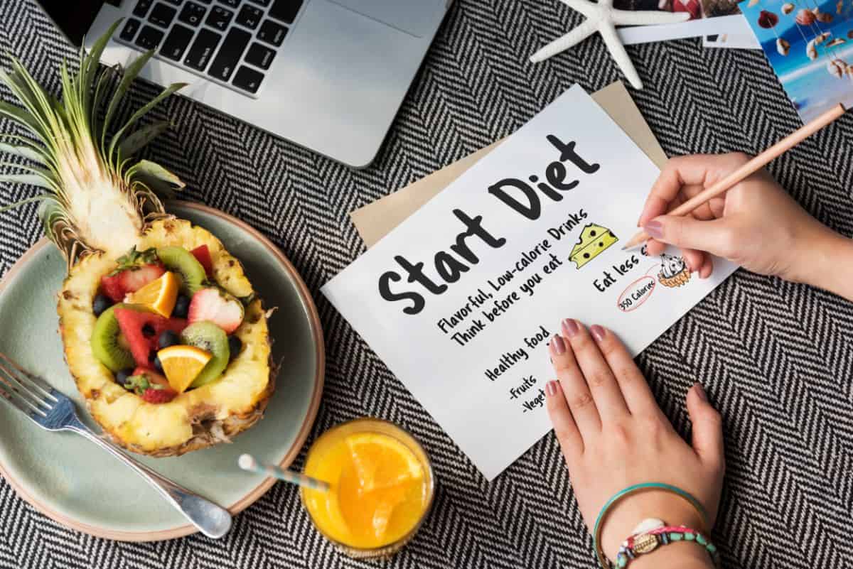 A person writing "Start Diet" on a piece of paper with a pencil. Next to the paper is a half pineapple filled with sliced fruits and a glass of orange juice. The scene also includes a laptop, a starfish, and a map on a zigzag patterned tablecloth.