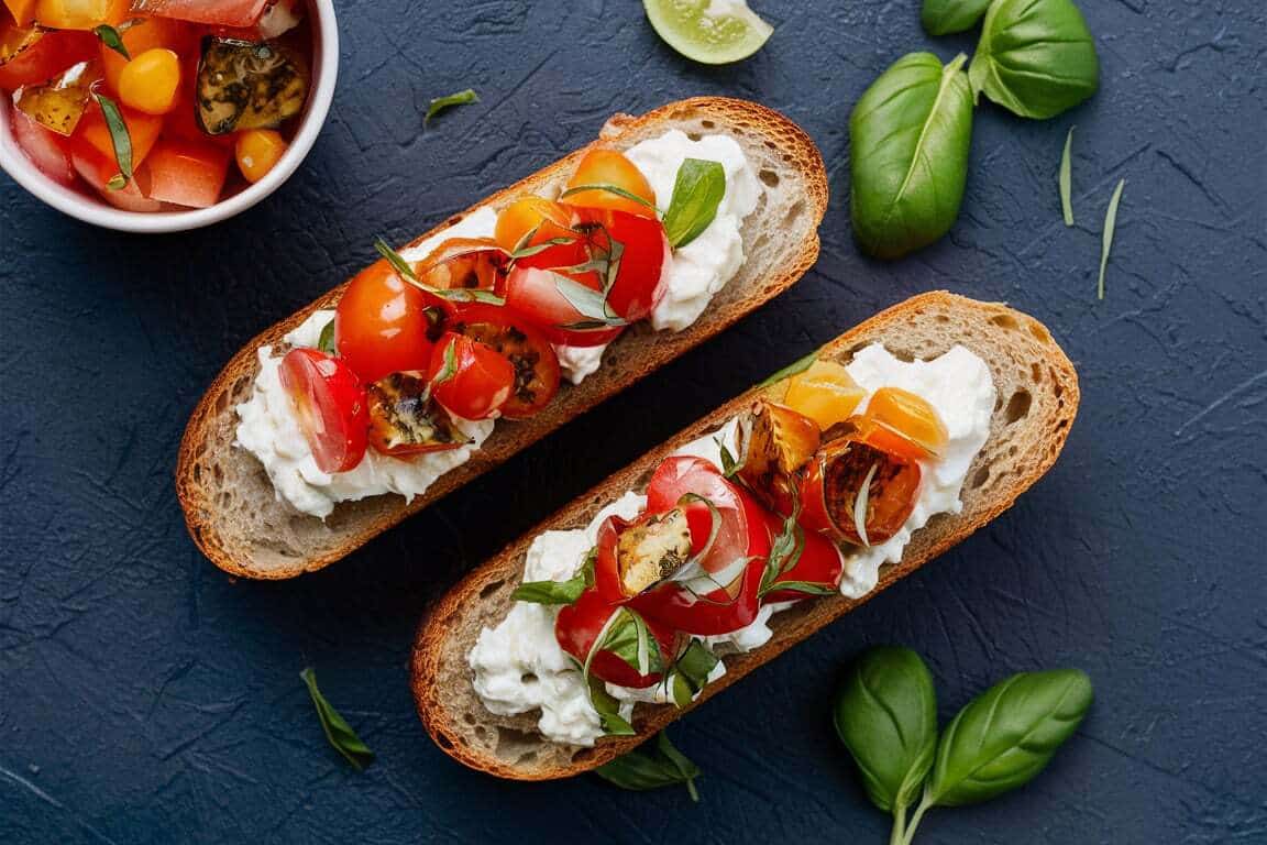 Two slices of crusty bread topped with cottage cheese, halved cherry tomatoes, fresh basil leaves, and a drizzle of olive oil. A small bowl with diced tomatoes and herbs is visible in the background, along with some loose basil leaves.