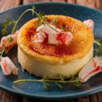 Best Crab Brulee Recipe A close-up image of a crab brûlée, showcasing a golden-brown caramelized top, garnished with crab meat and fresh herbs, served on a blue plate.