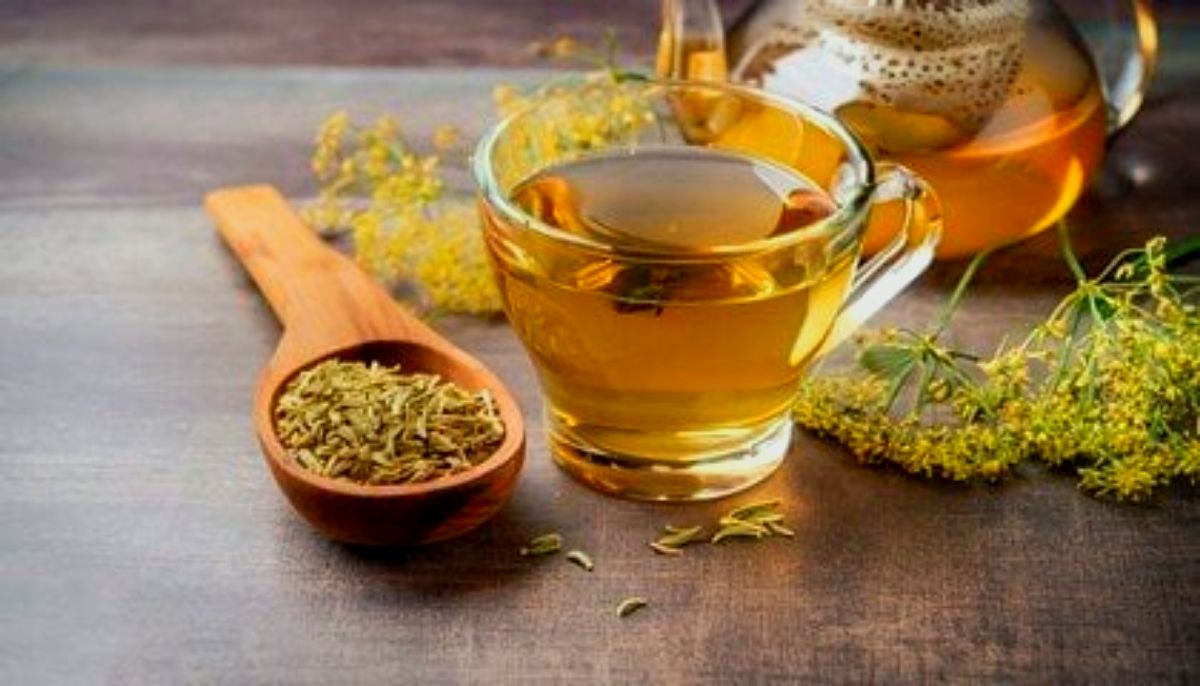 A glass cup filled with fennel tea, a wooden spoon containing fennel seeds, and a teapot in the background on a wooden surface with fresh fennel flowers.