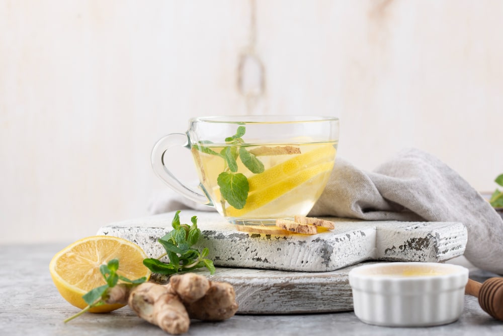 A glass cup of ginger mint tea with a lemon slice, placed on a rustic white wooden board. Fresh ginger, a half lemon, and mint leaves are arranged around the cup. A small white bowl with honey and a honey dipper are also visible.