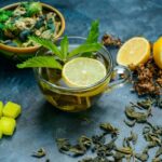 Herbal Tea Recipes A glass cup of herbal tea garnished with a lemon slice and fresh mint leaves. The background includes lemon halves, dried herbs, and green sugar cubes, all arranged on a dark blue surface.