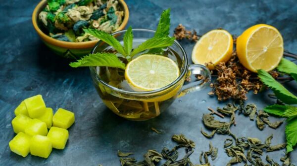 Herbal Tea Recipes A glass cup of herbal tea garnished with a lemon slice and fresh mint leaves. The background includes lemon halves, dried herbs, and green sugar cubes, all arranged on a dark blue surface.