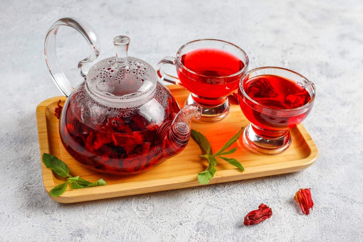 A clear glass teapot filled with hibiscus tea and two matching cups, placed on a wooden tray with fresh mint leaves as garnish.