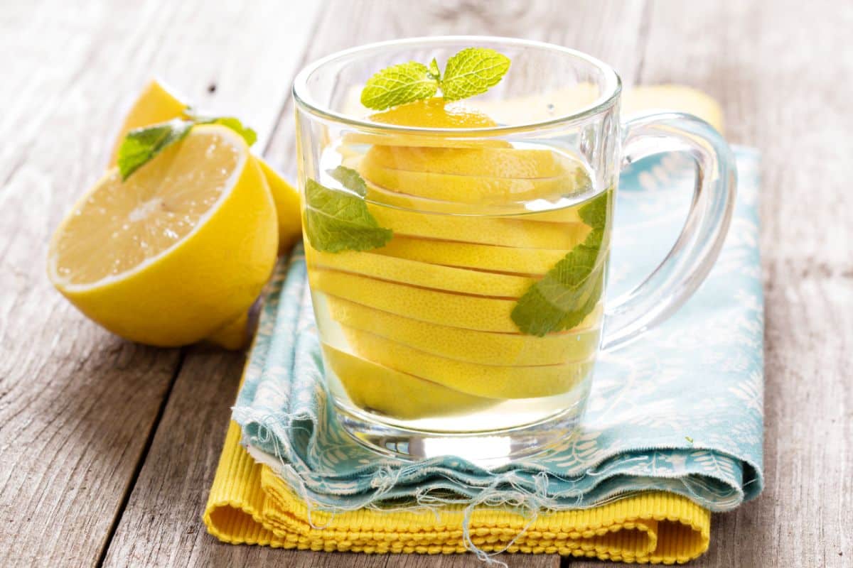 A glass cup of lemon balm tea with lemon slices and mint leaves, placed on a stack of colorful napkins. A halved lemon sits nearby on a rustic wooden surface.