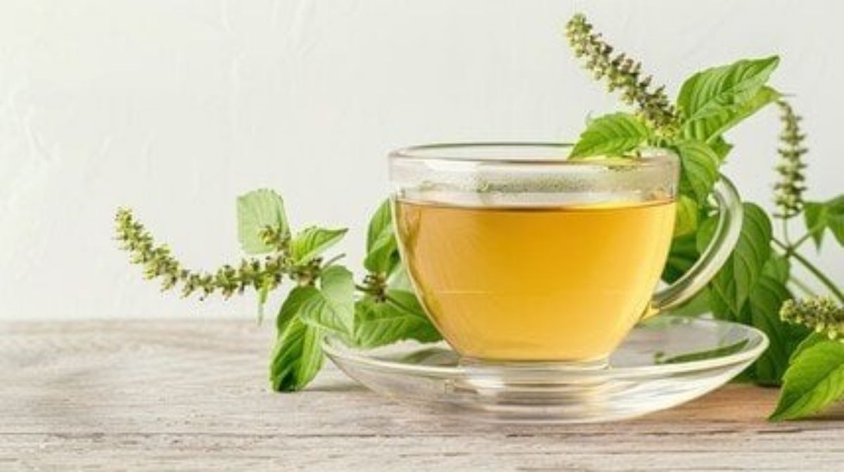 A glass cup filled with light brown tea sits on a clear saucer, with fresh green holy basil (tulsi) leaves and flowers placed beside it.