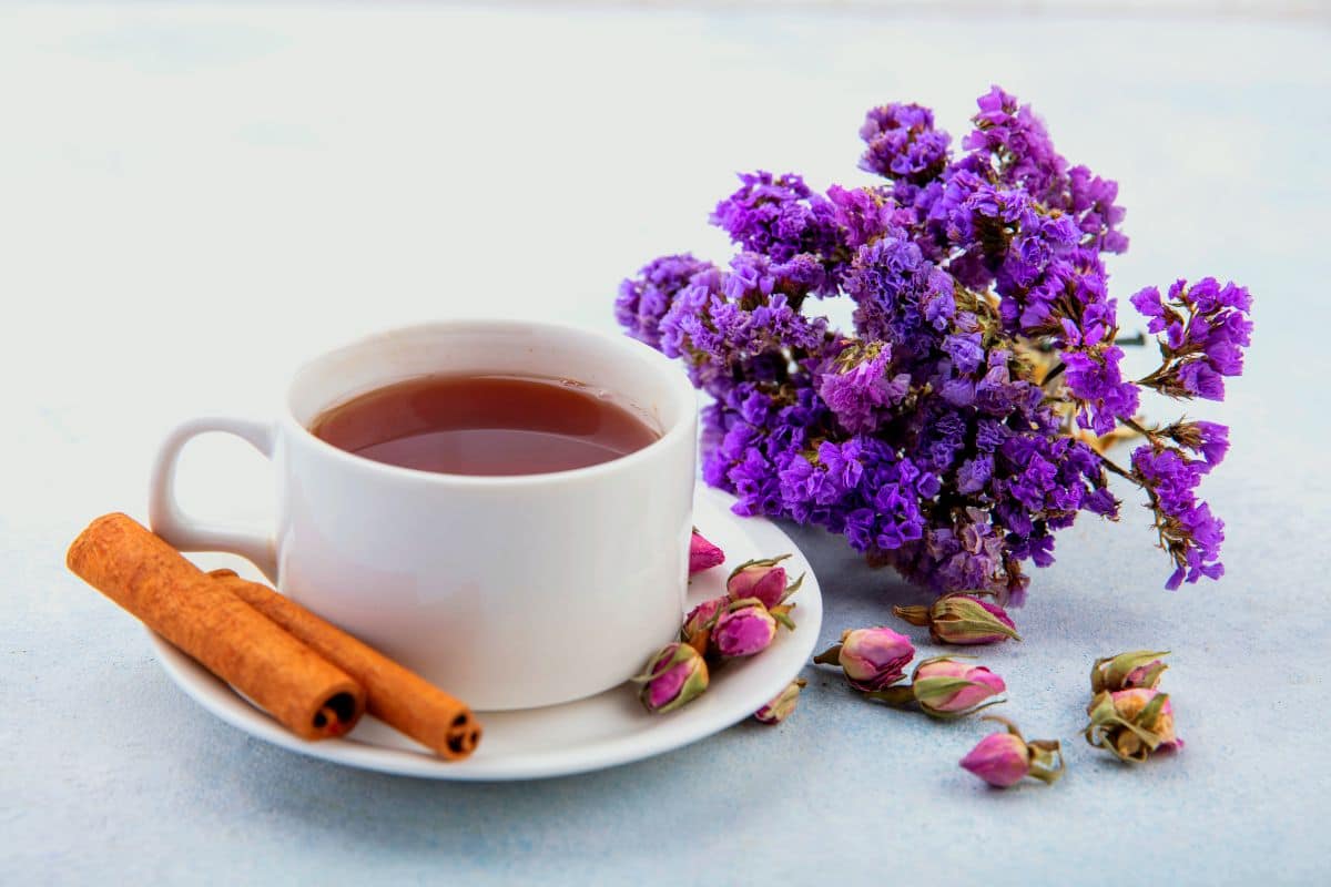 A white ceramic cup filled with chamomile lavender tea, accompanied by cinnamon sticks, dried rosebuds, and a bouquet of purple flowers on a light blue surface.