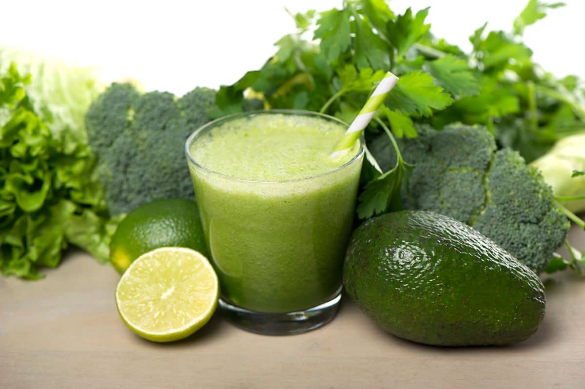 A glass of green avocado spinach smoothie with a striped straw, surrounded by fresh ingredients like avocado, lime, broccoli, spinach, and parsley.