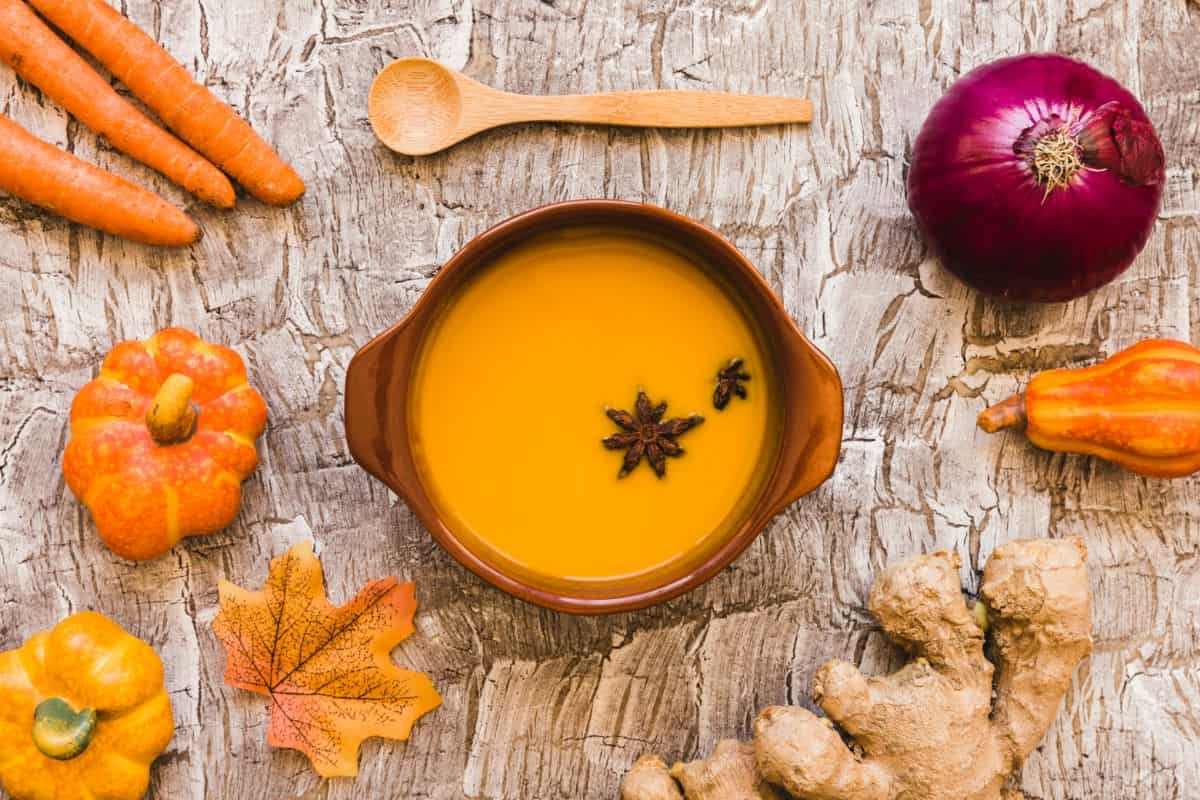 A brown ceramic bowl filled with carrot ginger soup, garnished with star anise, sitting on a rustic wooden surface. Surrounding the bowl are raw ingredients such as carrots, an onion, fresh ginger, small pumpkins, and a maple leaf. A wooden spoon is placed above the bowl.