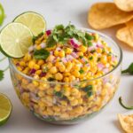 Chipotle Corn Salsa Recipe A bowl of colorful corn salsa garnished with lime slices and cilantro, surrounded by fresh limes, cilantro leaves, jalapeño peppers, and tortilla chips.