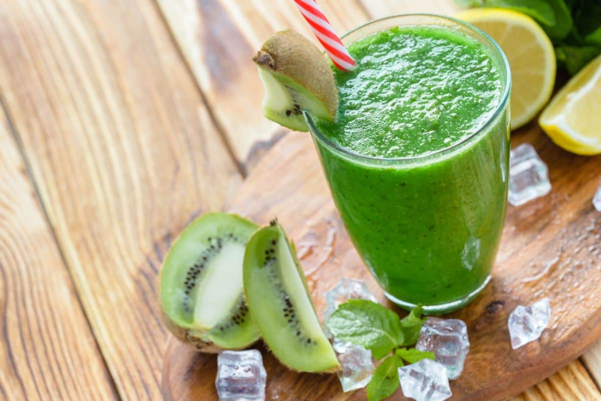 A glass of vibrant green smoothie garnished with a slice of kiwi, surrounded by fresh kiwi slices, lemon, mint leaves, and ice cubes on a wooden surface.