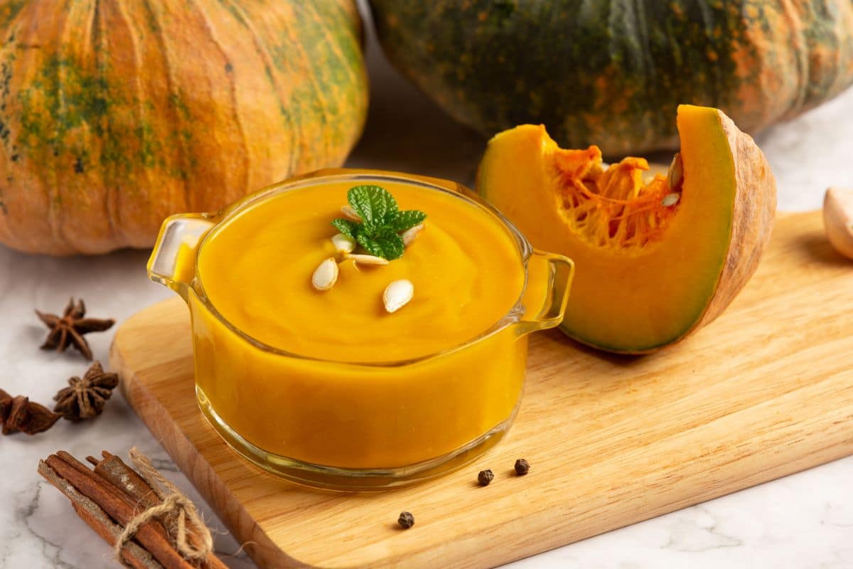 A glass bowl filled with creamy pumpkin soup garnished with pumpkin seeds and a sprig of mint. The bowl is placed on a wooden cutting board. In the background, there are halved pumpkins and spices such as cinnamon sticks and star anise.