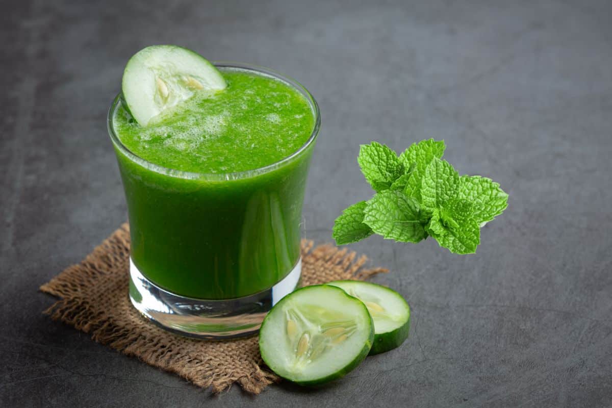 A refreshing green smoothie in a clear glass, garnished with a cucumber slice. Fresh mint leaves and cucumber slices are placed next to the glass on a textured surface.