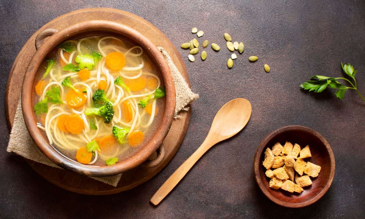 A bowl of vegetable broth with noodles, sliced carrots, and broccoli florets served in a rustic wooden bowl. A wooden spoon and a small wooden bowl filled with croutons are placed beside it on a dark, textured surface. Some pumpkin seeds and a sprig of parsley are scattered nearby for garnish.