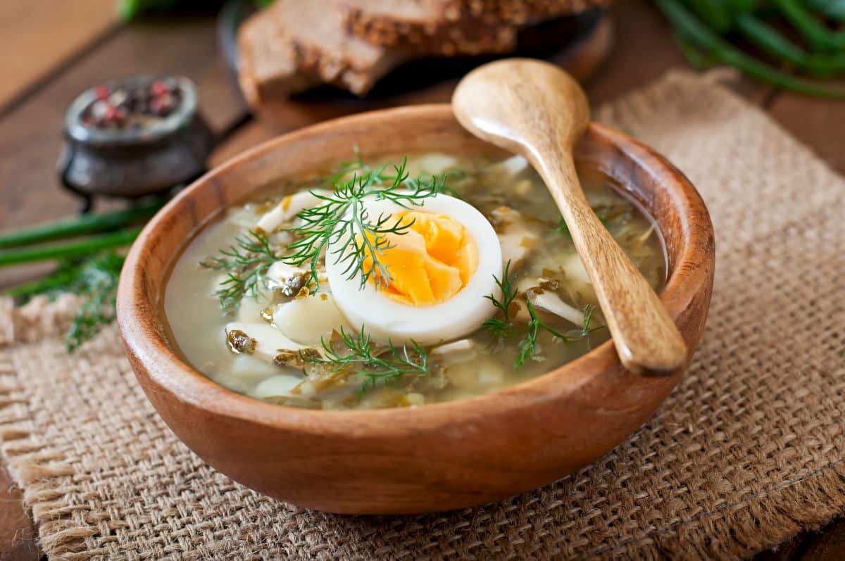 A wooden bowl filled with miso soup, garnished with half of a soft-boiled egg and fresh dill. A wooden spoon rests on the rim of the bowl, with slices of bread and a pepper grinder in the background.