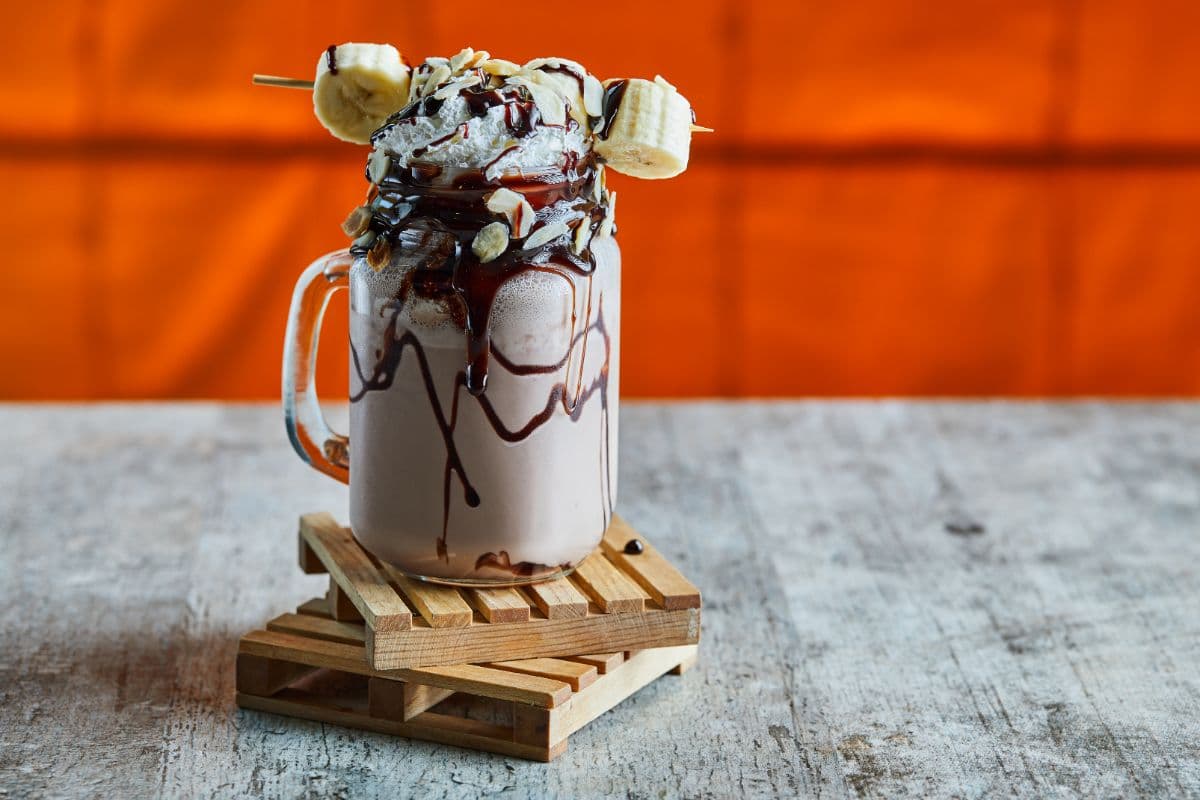 A mason jar filled with a thick chocolate smoothie, topped with whipped cream, chocolate drizzle, and banana slices. The smoothie is garnished with sliced almonds and a skewer of banana pieces, served on a small wooden coaster. The background is a vibrant orange, contrasting with the rustic wooden surface below.