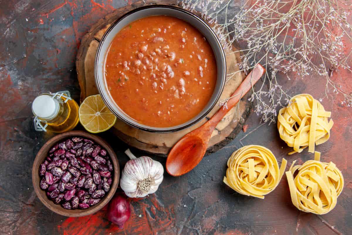 A bowl of hearty tomato soup with beans, surrounded by a wooden spoon, bundles of uncooked pasta, garlic, dried beans, and a bottle of olive oil on a rustic wooden board.