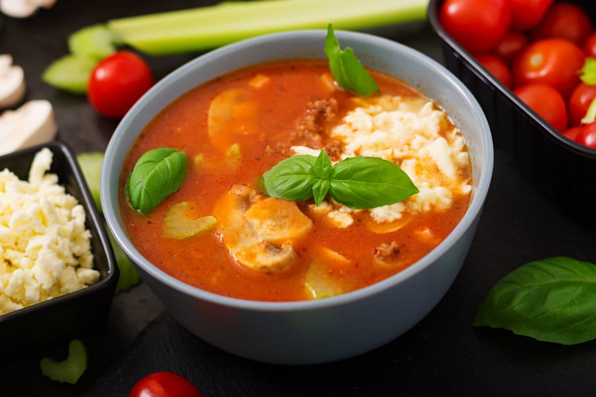 A bowl of tomato basil soup topped with fresh basil leaves and a sprinkle of cheese. The soup is vibrant red and is served in a light gray bowl. Surrounding the bowl are ingredients including cherry tomatoes, celery sticks, and a dish of shredded cheese.