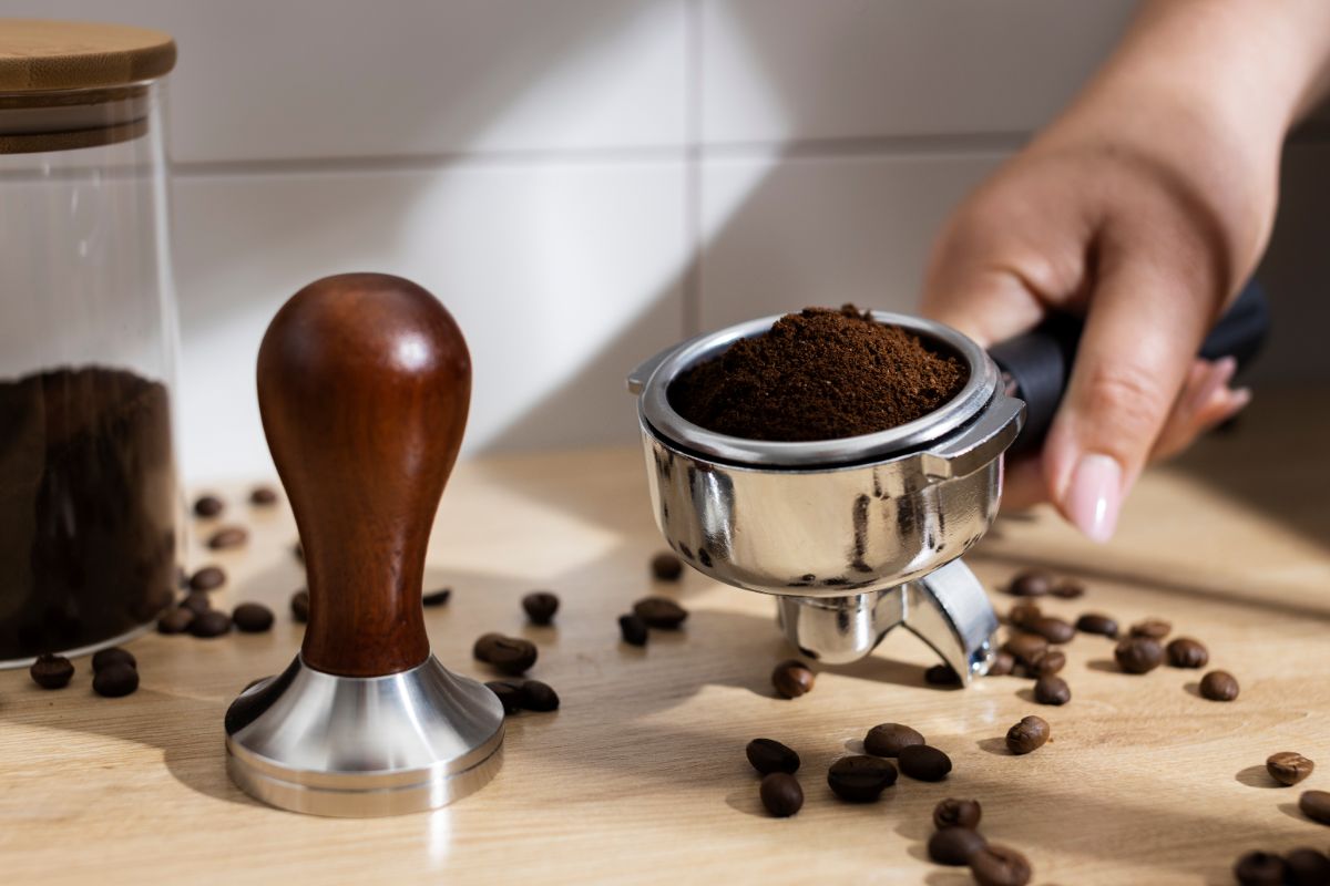 A hand holding a portafilter filled with freshly ground coffee beans, with scattered whole coffee beans around on a wooden surface. A wooden coffee tamper is positioned next to the portafilter. In the background, there is a jar filled with coffee grounds. The setting is well-lit, creating a cozy coffee preparation scene.