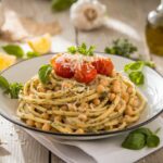 Chickpea Pasta A plate of spaghetti topped with roasted cherry tomatoes, chickpeas, and grated cheese, garnished with fresh basil, sitting on a rustic wooden table alongside lemon wedges, garlic, and olive oil.