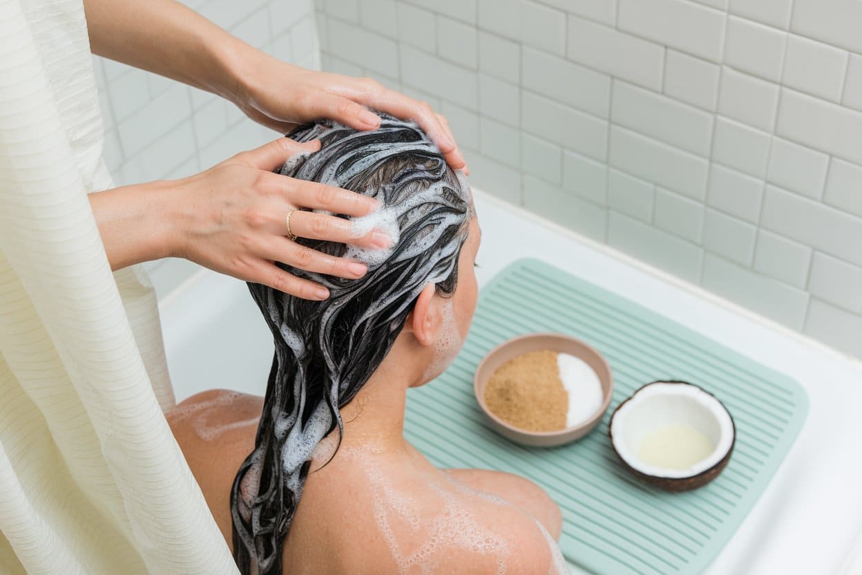 A person seated in a bathtub getting their hair washed with foamy shampoo as another person massages their scalp.