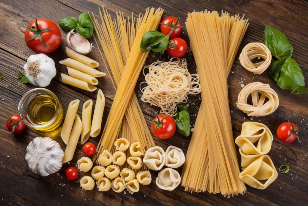 Different Types Of Pasta A Variety Of Uncooked Pasta Types Displayed On A Wooden Table Alongside Ingredients Like Tomatoes, Garlic, Basil, And Olive Oil. The Pasta Includes Spaghetti, Penne, Tortellini, And Other Shapes, Neatly Arranged In A Visually Appealing Manner.