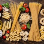 Different Types Of Pasta A Variety Of Uncooked Pasta Types Displayed On A Wooden Table Alongside Ingredients Like Tomatoes, Garlic, Basil, And Olive Oil. The Pasta Includes Spaghetti, Penne, Tortellini, And Other Shapes, Neatly Arranged In A Visually Appealing Manner.