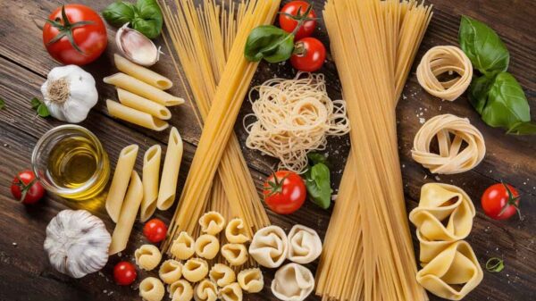 Different Types of Pasta A variety of uncooked pasta types displayed on a wooden table alongside ingredients like tomatoes, garlic, basil, and olive oil. The pasta includes spaghetti, penne, tortellini, and other shapes, neatly arranged in a visually appealing manner.