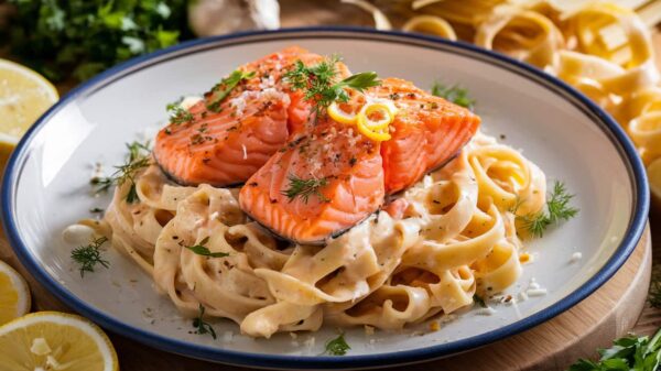 Salmon Pasta A plate of creamy fettuccine pasta topped with two fillets of grilled salmon, garnished with fresh herbs and lemon zest, served with slices of lemon on the side.