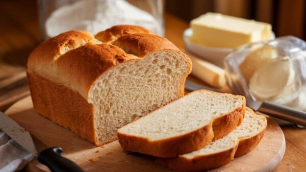 Sandwich Bread Recipe A Freshly Baked Loaf Of Homemade Sandwich Bread On A Wooden Cutting Board, With A Few Slices Cut. The Bread Has A Golden-Brown Crust And Soft, Fluffy Interior. Surrounding The Bread Are Ingredients Like Butter And Flour, Creating A Cozy Kitchen Setting.